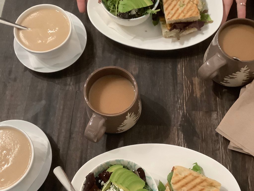 A top-down view of a lunch meeting in the Cowichan Valley featuring coffee mugs, paninis, and avocado salads on a dark wood table.